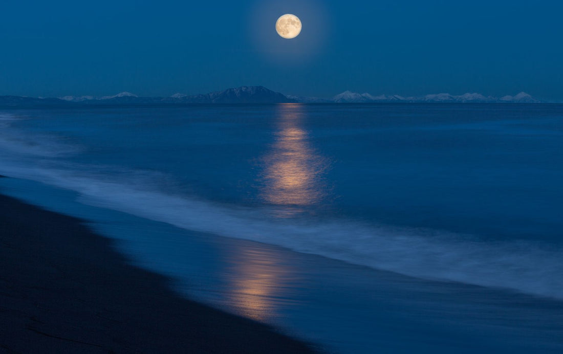A full moon rising over the ocean, its light reflecting across the water toward a Hawaiʻi beach — an image of Hina, goddess of the moon