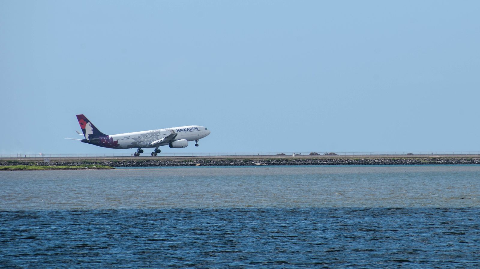 A Hawaiian Airlines jet taking off over the water near a Hawaiʻi airport runway