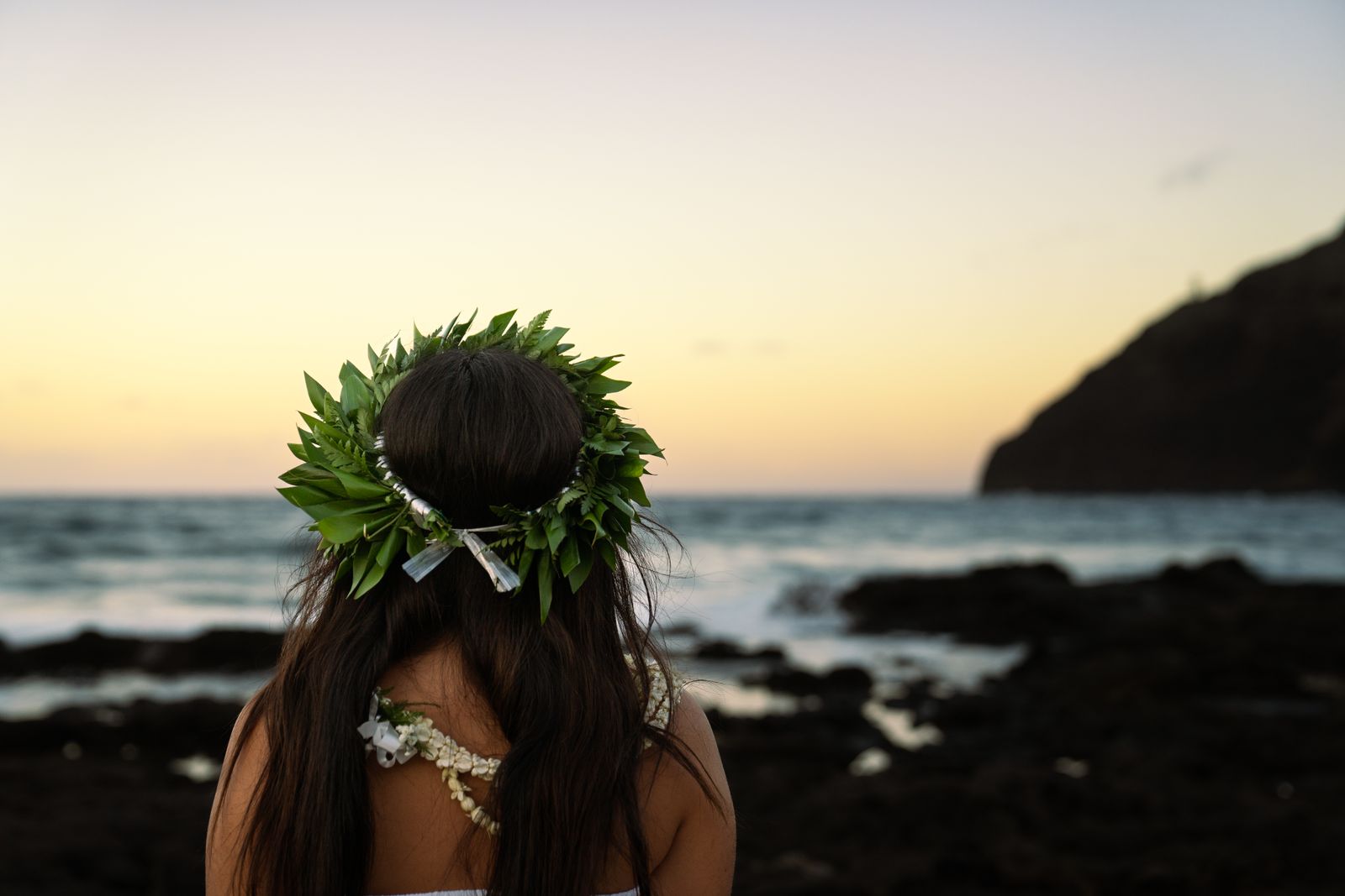 Woman wearing a traditional haku lei crown and lei necklace at the Hawaiian coastline during sunset, evoking blessing and ceremony.