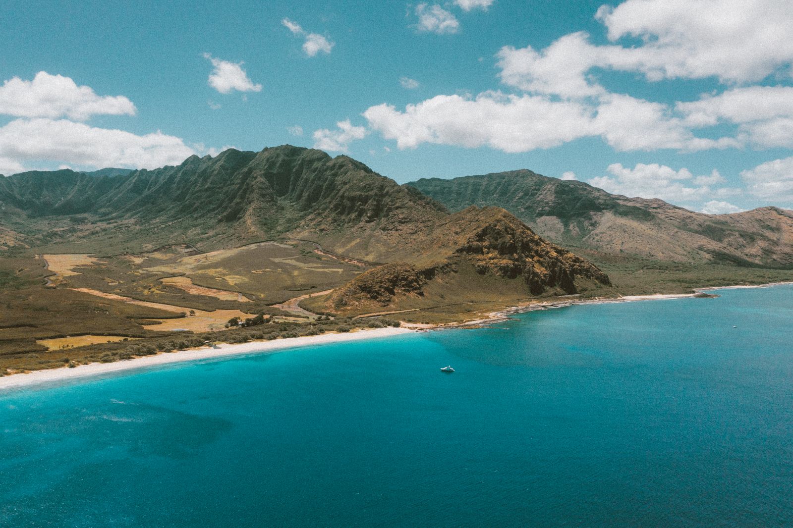 Aerial view of a Hawaiian island coastline with green mountains meeting a white-sand beach and turquoise ocean in Hawaiʻi