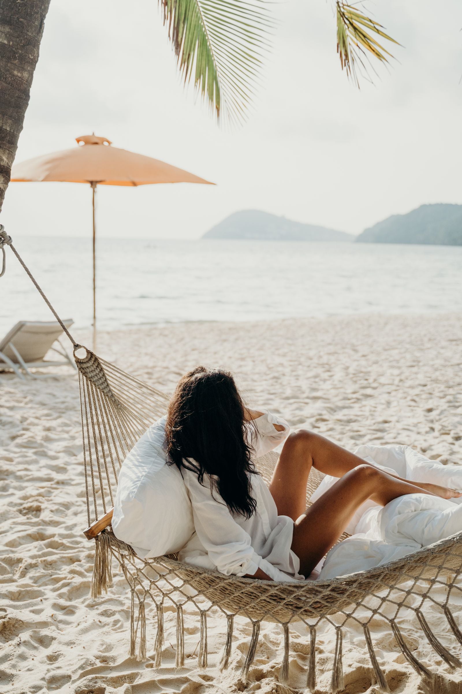 A woman relaxing barefoot in a beach hammock under a palm tree beside a calm tropical bay, living the aloha lifestyle in Hawaiʻi